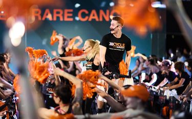 A man and a woman wearing a Cycle for Survival t-shirts smiles on stationary bikes surrounded by others riding stationary bikes and waving orange pom-poms.