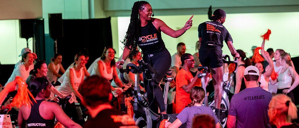 A cycling instructor in a black Cycle for Survival tank top on a podium in the center of a room, surrounded by people on stationary bikes